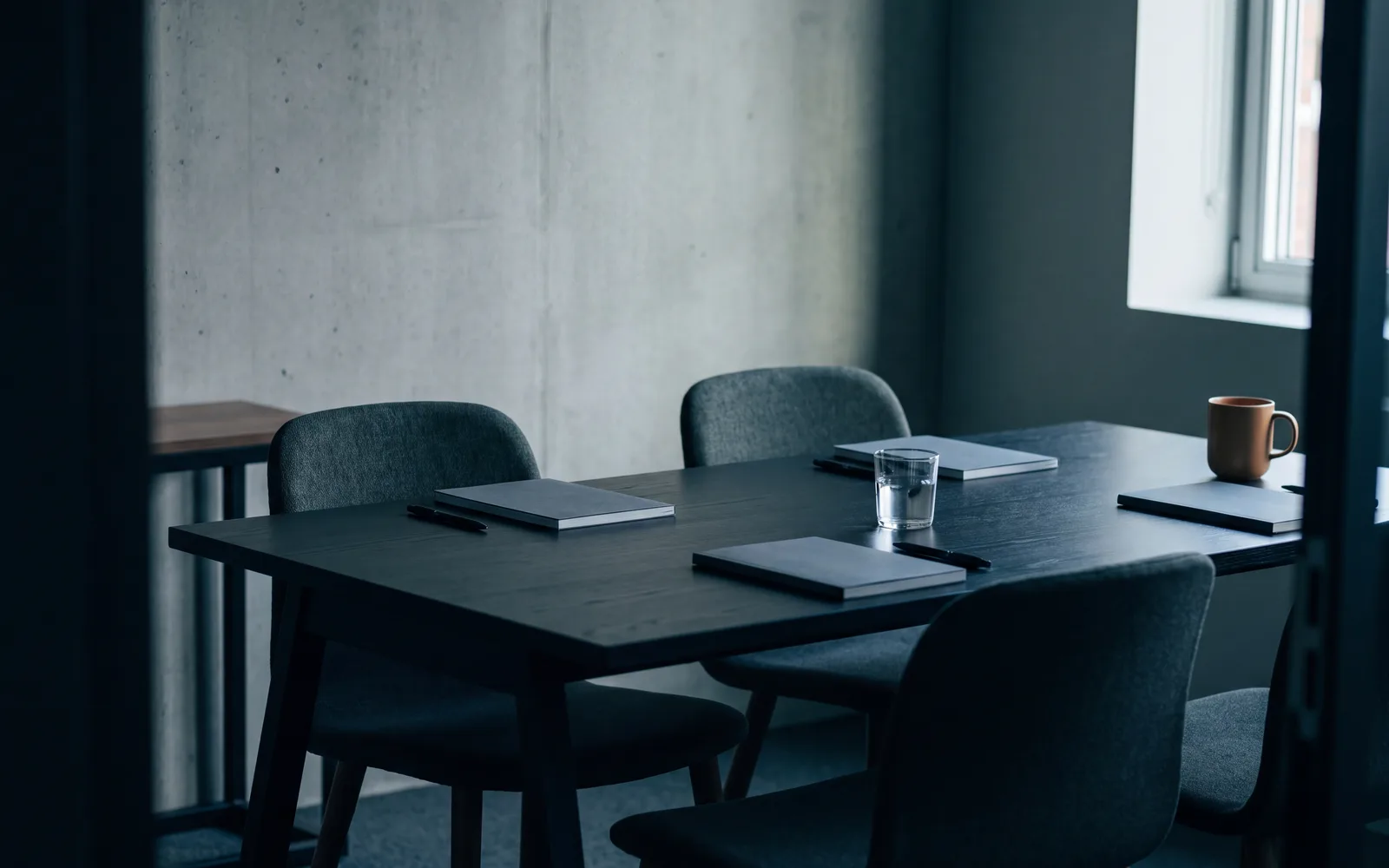 A work table with chairs around it, notebooks and pens, a glass of water and a cup, light from a window